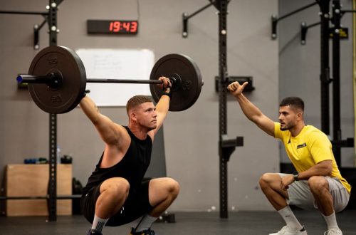 Athlete performing weightlifting exercise under trainer's guidance in a gym setting.