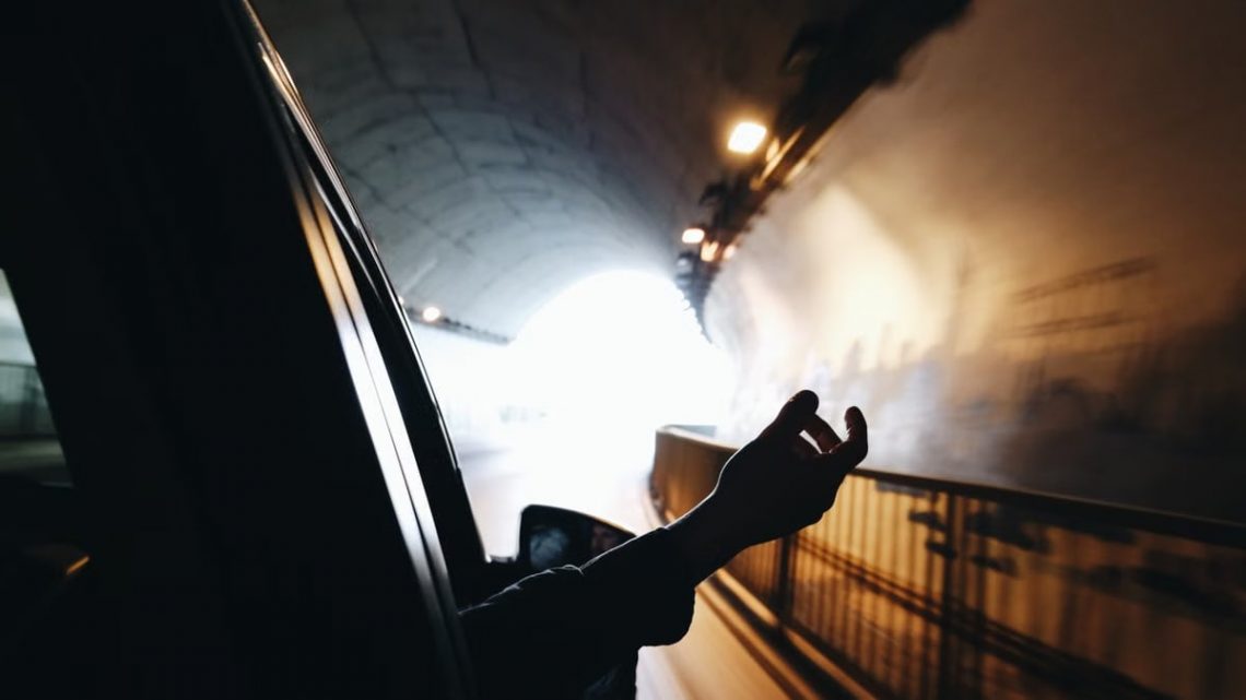 A hand extends out of a car window while driving through a brightly lit tunnel.